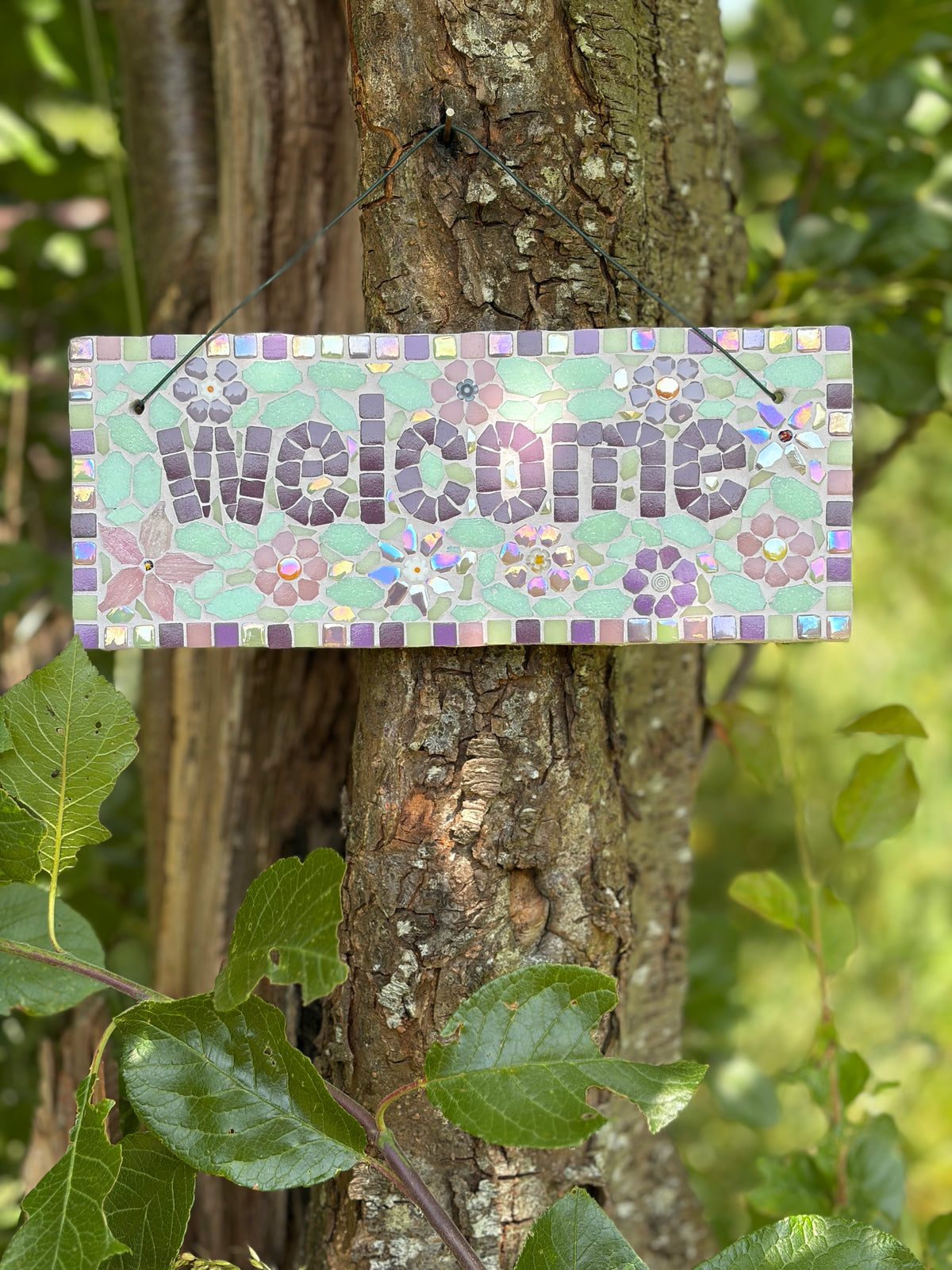 Handcrafted mosaic welcome sign in soft purples and mint greens with floral motifs, hung outdoors
