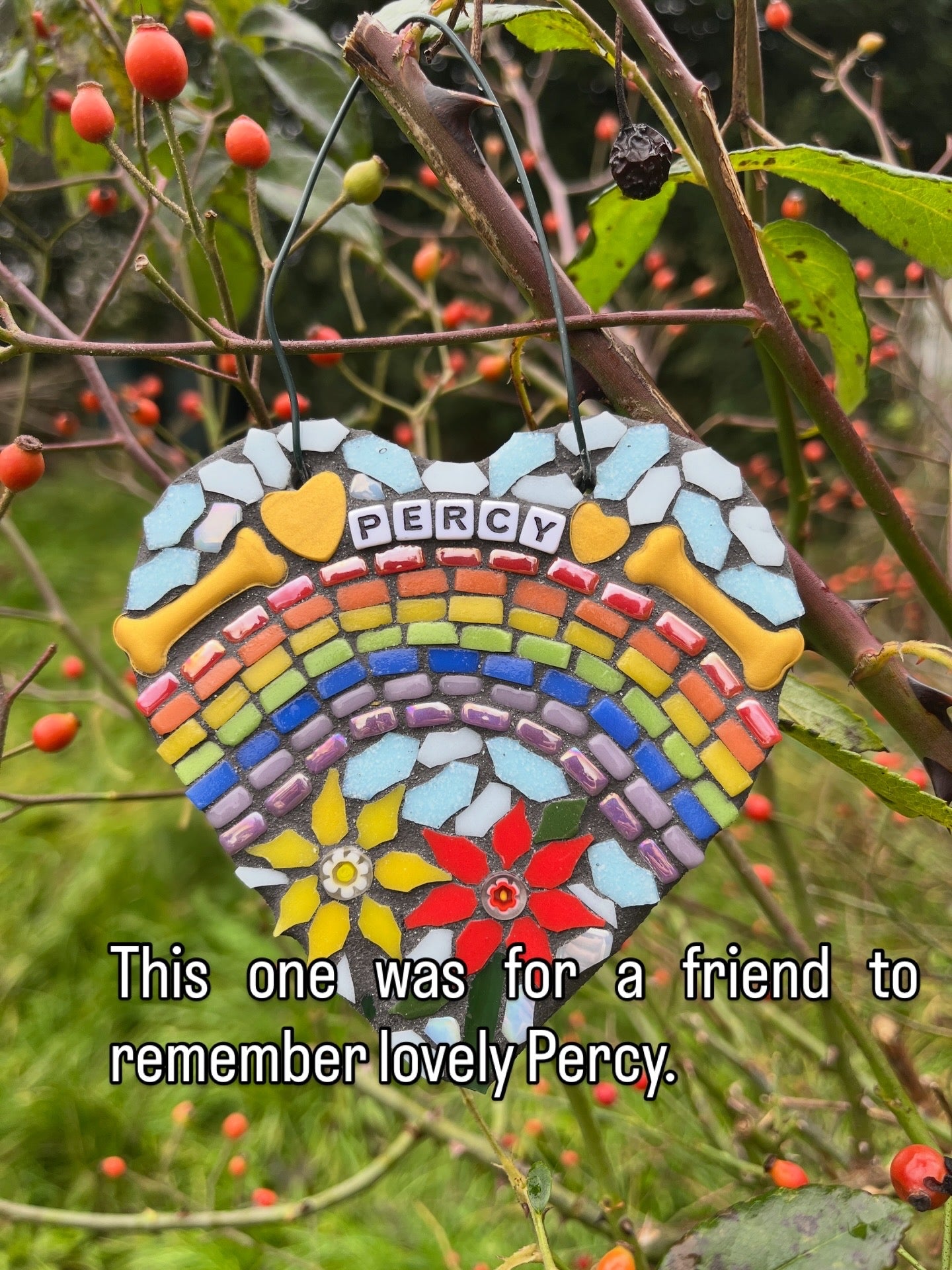 Personalised dog memorial mosaic heart with rainbow arch, dog's name in letter tiles, golden dog bones and handmade glass flowers, hung outdoors