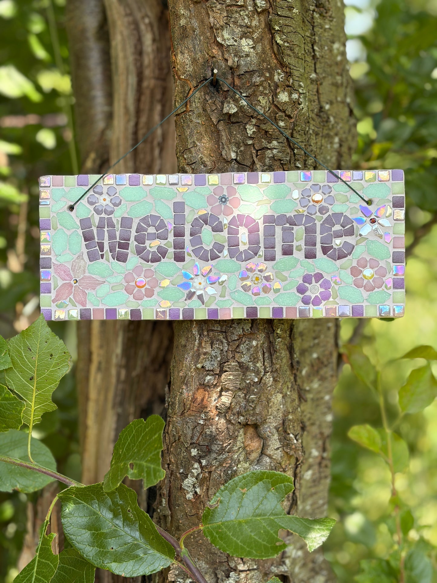 Close-up of floral mosaic welcome sign showing purple and mint tile detail, hung on a tree
