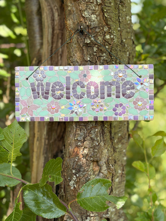 Close-up of floral mosaic welcome sign showing purple and mint tile detail, hung on a tree