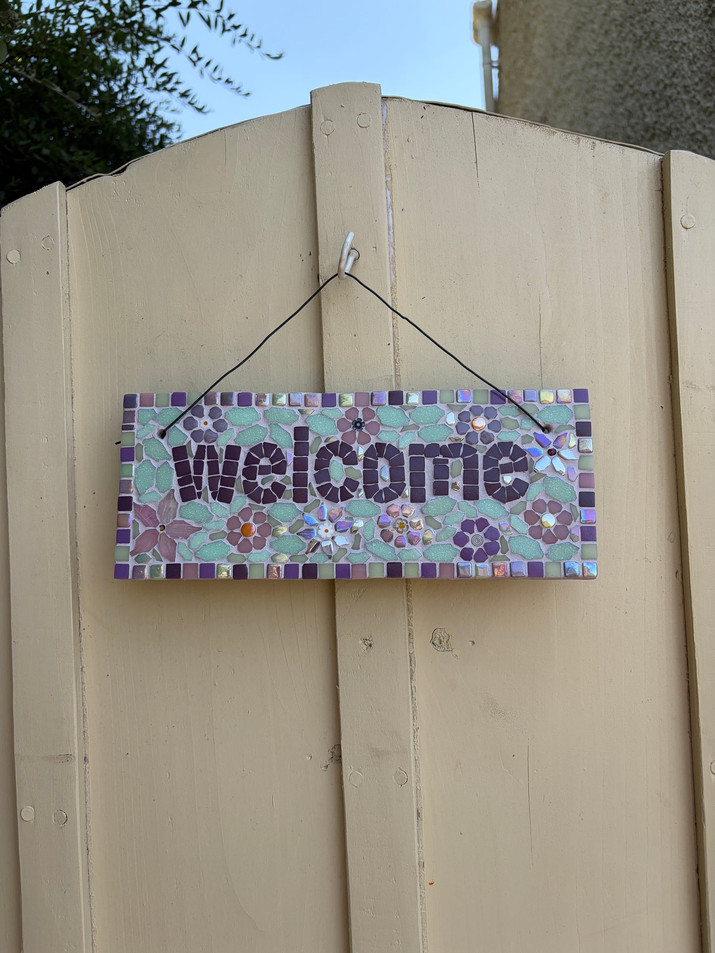 Handcrafted floral mosaic welcome sign on slate with purple border, hung on a cream garden gate