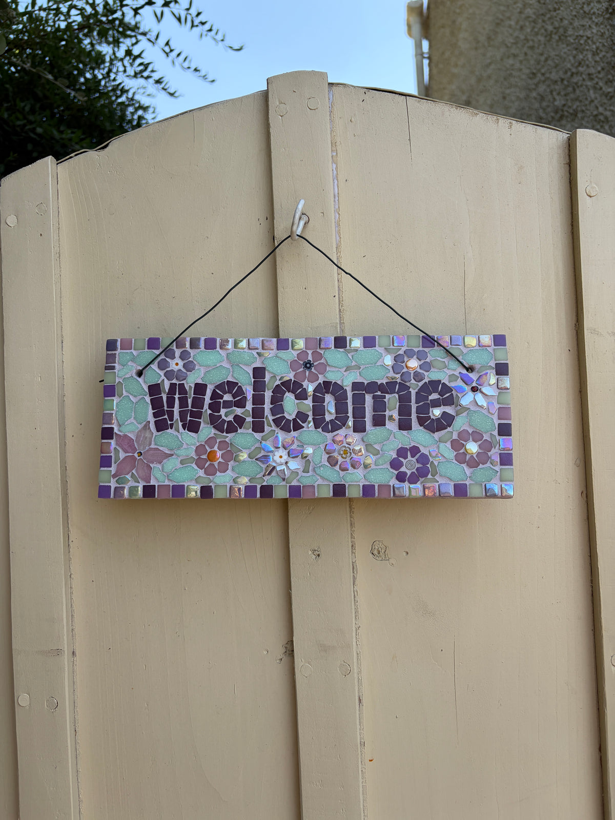 Handcrafted floral mosaic welcome sign on slate with purple border, hung on a cream garden gate