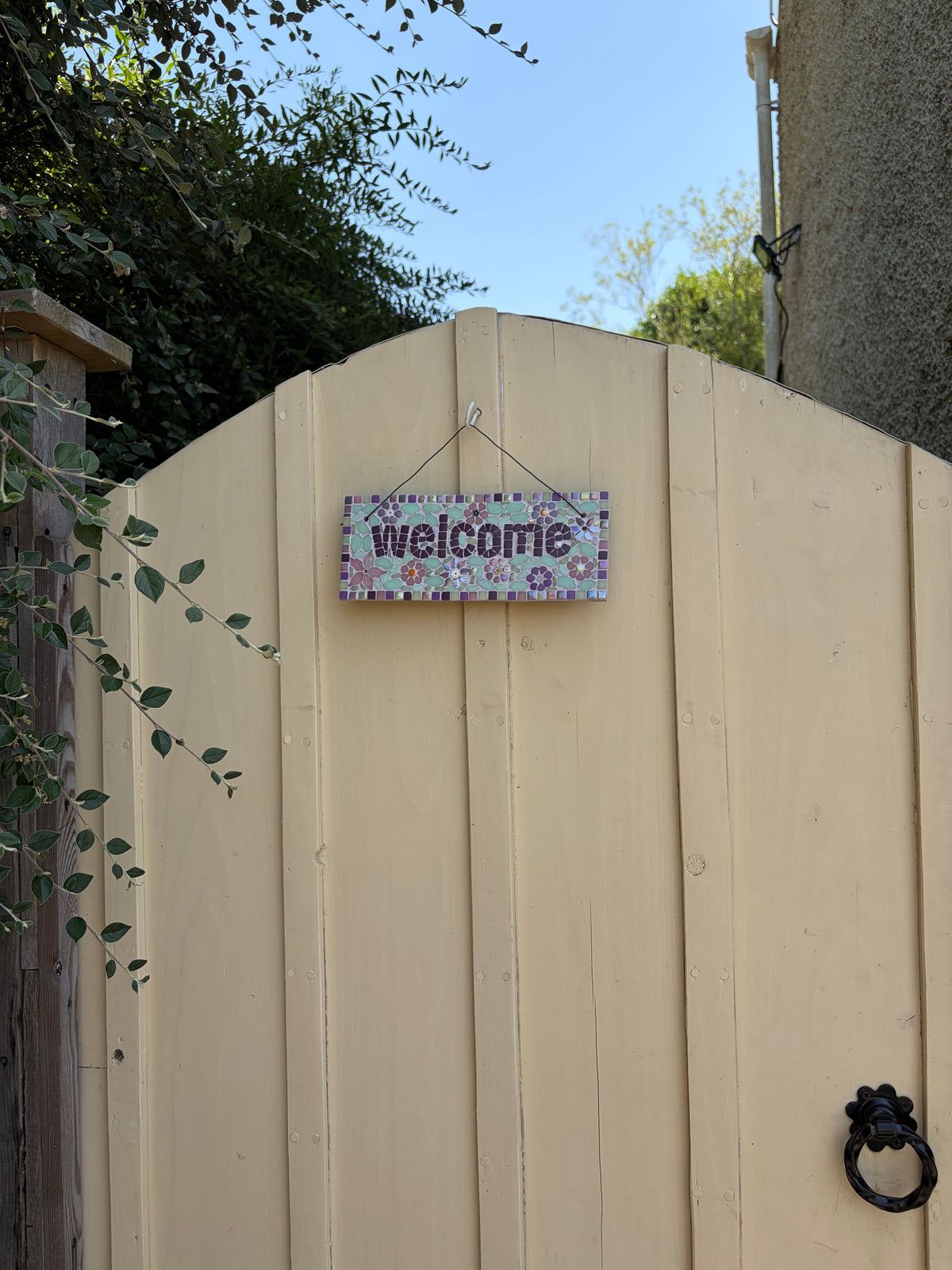 Floral mosaic welcome sign hung on a cream garden gate, showing full context
