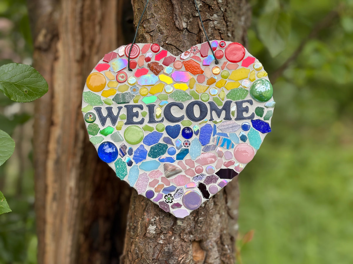 Rainbow mosaic heart welcome sign on slate hung on a tree, showing full piece from a slight distance