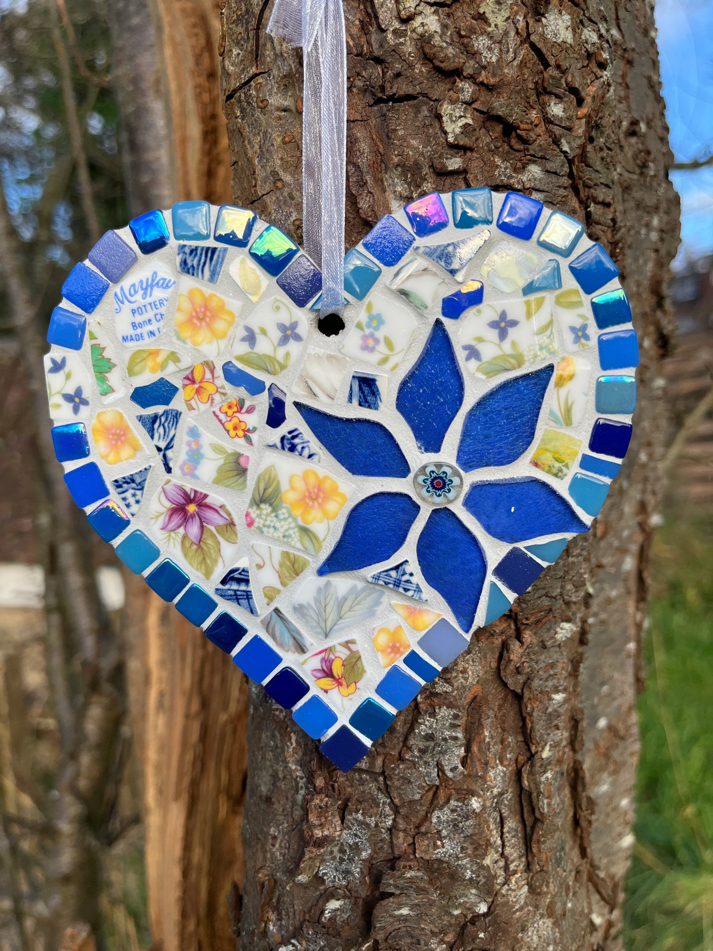 Handcrafted mosaic heart using vintage Mayfair Pottery bone china with yellow and purple wildflowers, deep cobalt blue glass flower centrepiece and blue tile border with iridescent accents, hung on a tree