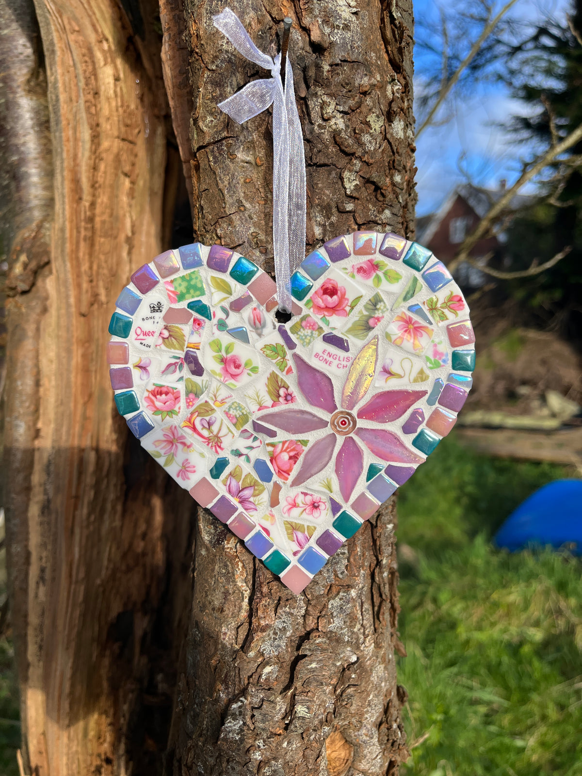 Handcrafted mosaic heart using vintage Queen's bone china with pink roses, iridescent purple glass flower centrepiece and jewel-tone iridescent tile border, hung on a tree