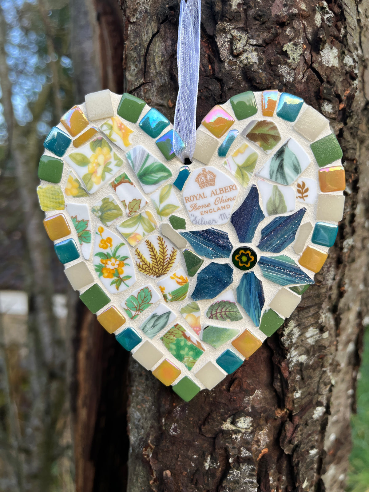 Handcrafted mosaic heart using Royal Albert Silver Maple bone china with teal, amber and sage green tile border and blue glass flower centrepiece, hung on a tree