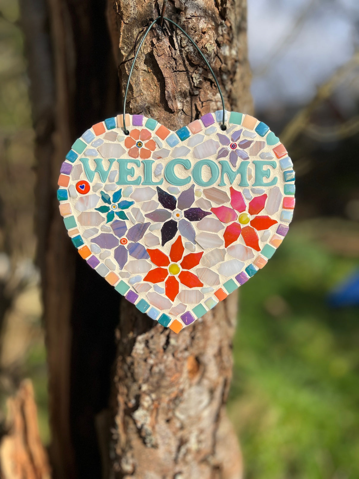 Handcrafted mosaic floral welcome heart on slate with teal ceramic WELCOME lettering, colourful flower mosaic and rainbow tile border, hung on a tree