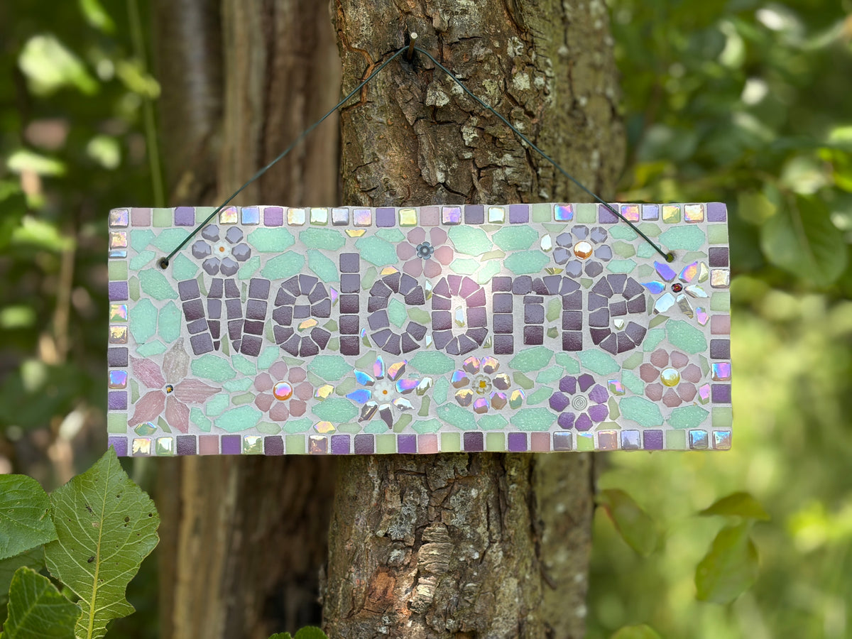 Mosaic welcome sign with iridescent tiles and purple floral design, hung on a tree