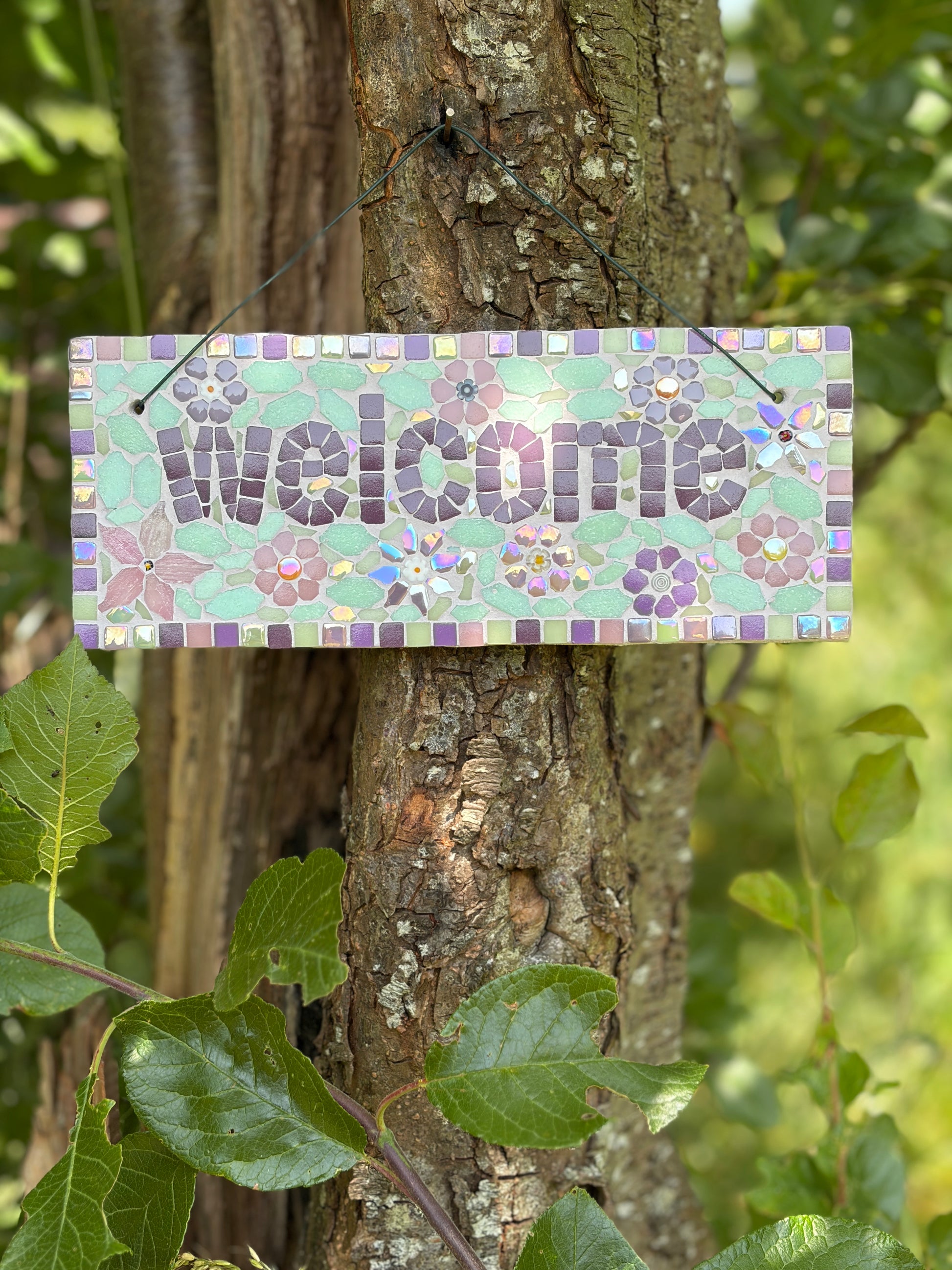 Handcrafted mosaic welcome sign in soft purples and mint greens with floral motifs, hung outdoors