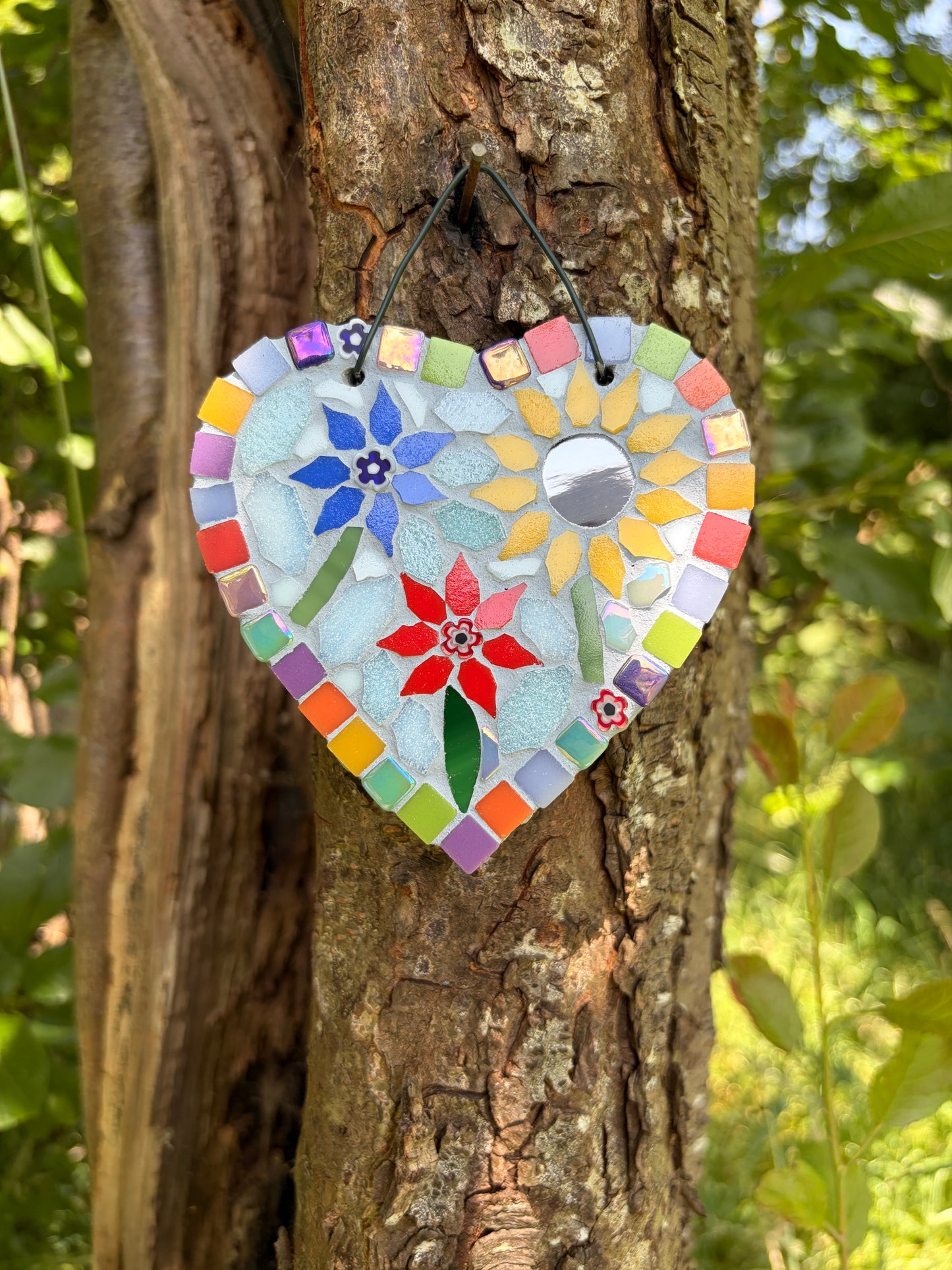 Handcrafted floral mosaic heart decoration on slate with rainbow border and cord hanging loop, photographed on a tree